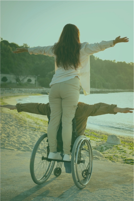 Eine Frau steht lachend mit ausgebreiteten Armen auf einem Rollstuhl am Strand